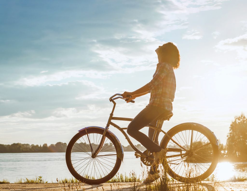 Side View Of Woman Riding Bicycle By Lake Against Sky At Sunset