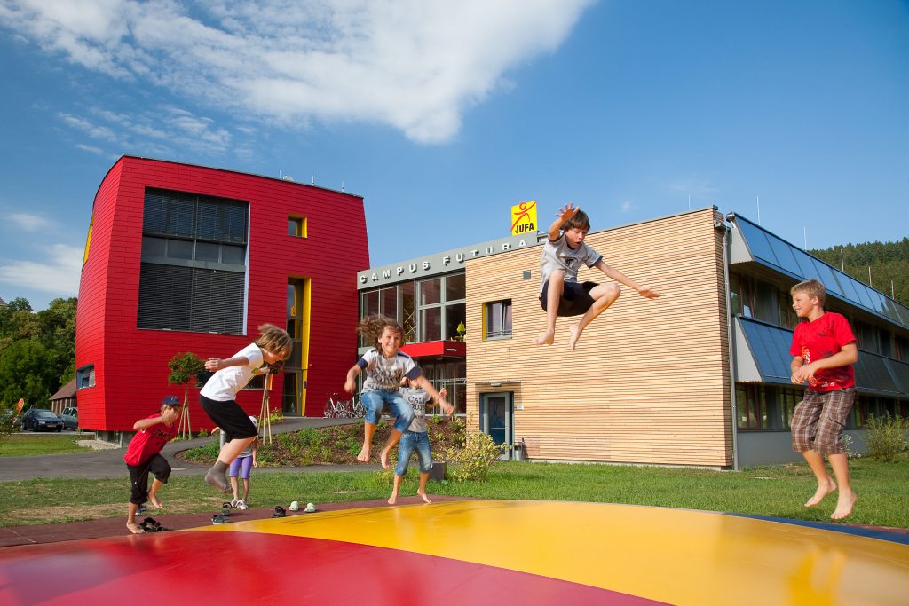 Sie sehen eine Außenansicht vom JUFA Hotel Bleiburg/Pliberk – Sport-Resort mit Kindern im Sommer. JUFA Hotels bietet Ihnen den Ort für erfolgreiches Training in ungezwungener Atmosphäre für Vereine und Teams.