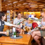 Sie sehen eine Familie im JUFA Alpenhotel Saalbach****, die von einer Kellnerin Getränke bekommt. JUFA Hotels bietet erholsamen Familienurlaub und einen unvergesslichen Winter- und Wanderurlaub.