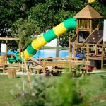 Sie sehnen den Spielplatz mit Holzgeräten und einer Rutsche und Bäumen im Hintergrund beim JUFA Hotel Neutal Landerlebnis. Die Zeit spielend und abwechslungsreich verbringen im JUFA Hotel Neutal - Landerlebnis. Der Ort für erlebnisreichen Natururlaub für die ganze Familie.