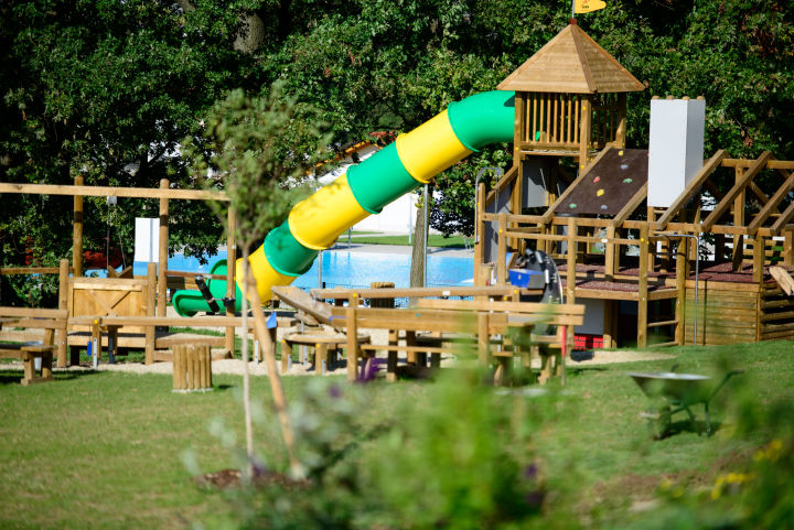 Sie sehnen den Spielplatz mit Holzgeräten und einer Rutsche und Bäumen im Hintergrund beim JUFA Hotel Neutal Landerlebnis. Die Zeit spielend und abwechslungsreich verbringen im JUFA Hotel Neutal - Landerlebnis. Der Ort für erlebnisreichen Natururlaub für die ganze Familie.