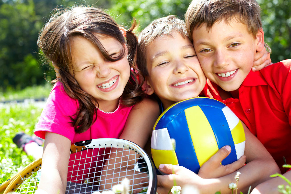 Three,Children,With,Sports,Equipment,Embracing,,Looking,At,Camera,And